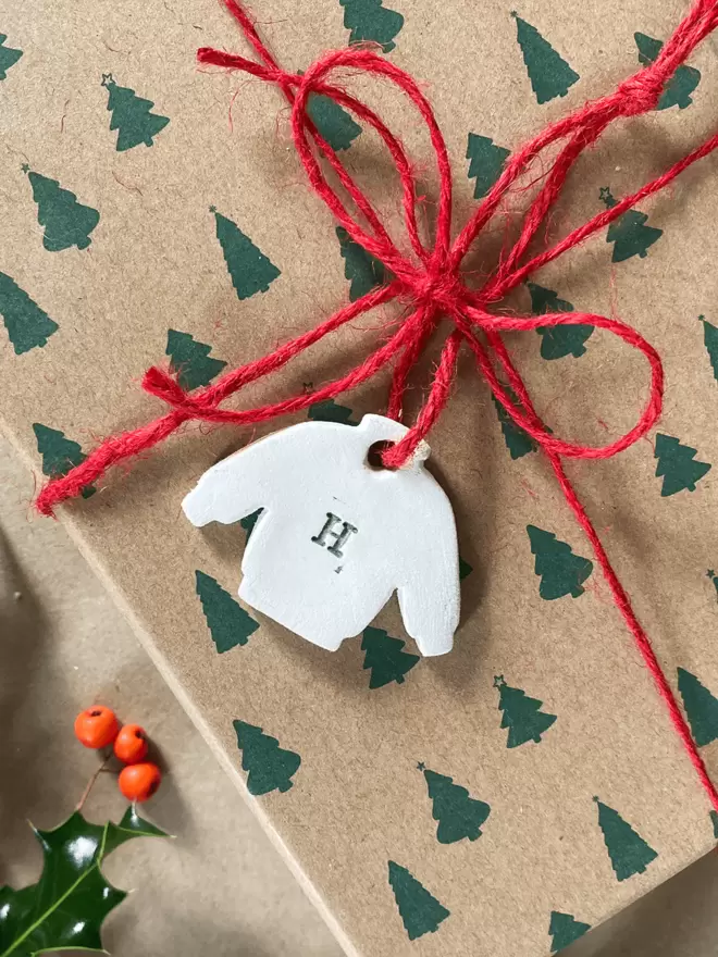 Small clay jumper tag with a 'H' on attached with red twine to a christmas present wrapped in brown paper with green christmas trees on. There is a sprig of holly in the background.