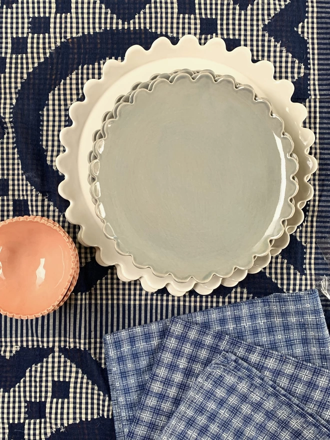 white daisy scallop dinner plate stcked with taupe grey daisy side plates on a checkered cloth with blue checkered napkins