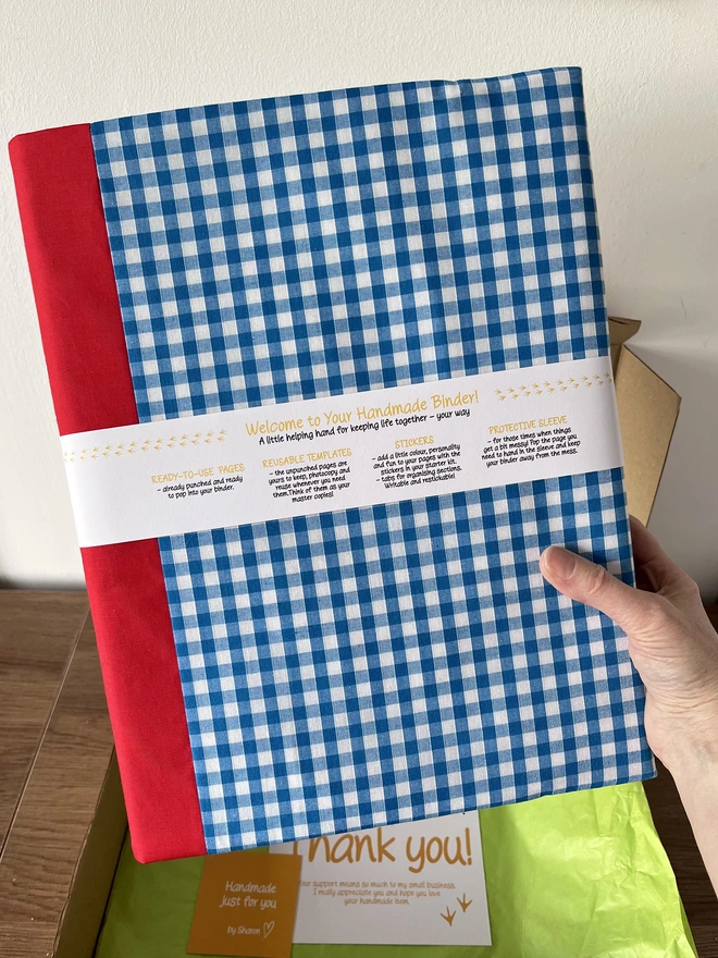 Close-up of hand holding up a ring binder with blue gingham and red fabric cover. It has a white belly band packing around it saying 'Welcome to Your Handmade Binder!' with instructions beneath on how to use the template pages and stickers of the planner starter kit included. Open kraft postal box with lime tissue & thank you card inside in background.