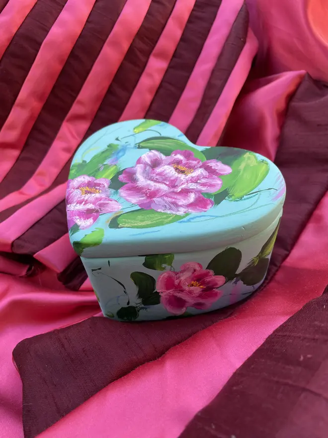 hand painted ceramic heart shaped keepsake box background painted In a light blue with pink and white peony flowers.