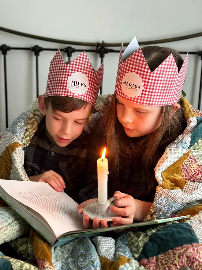 Two children wearing red gingham personalised Christmas crowns are wrapped in a cosy patchwork quilt reading a book together by candlelight.
