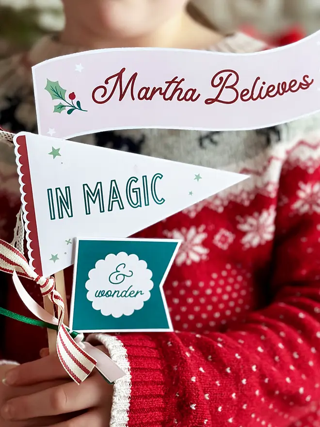 A child wearing a traditional red Christmas jumper holds three personalised Christmas flags made from cardboard. Each flag is attached to a wooden stick with ribbons. The flags read "Martha Believes in magic & wonder".