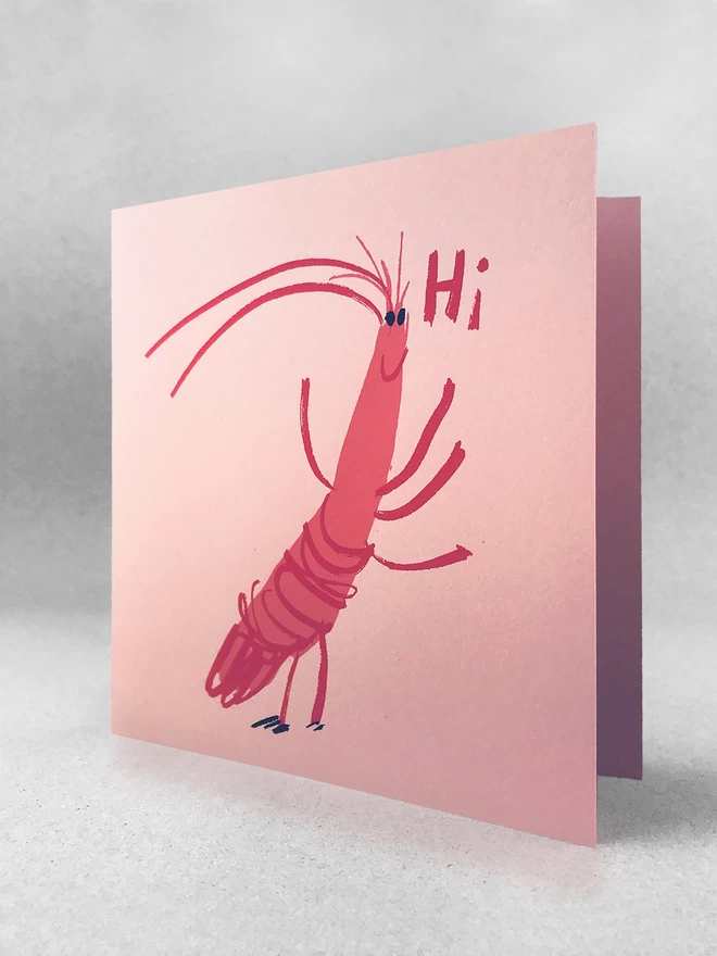A happy cartoon prawn says Hi on this jolly upbeat pink card, printed in two pink inks with black eyes. It is angled to see the blank card inside, and the card is stood on a light grey background. 