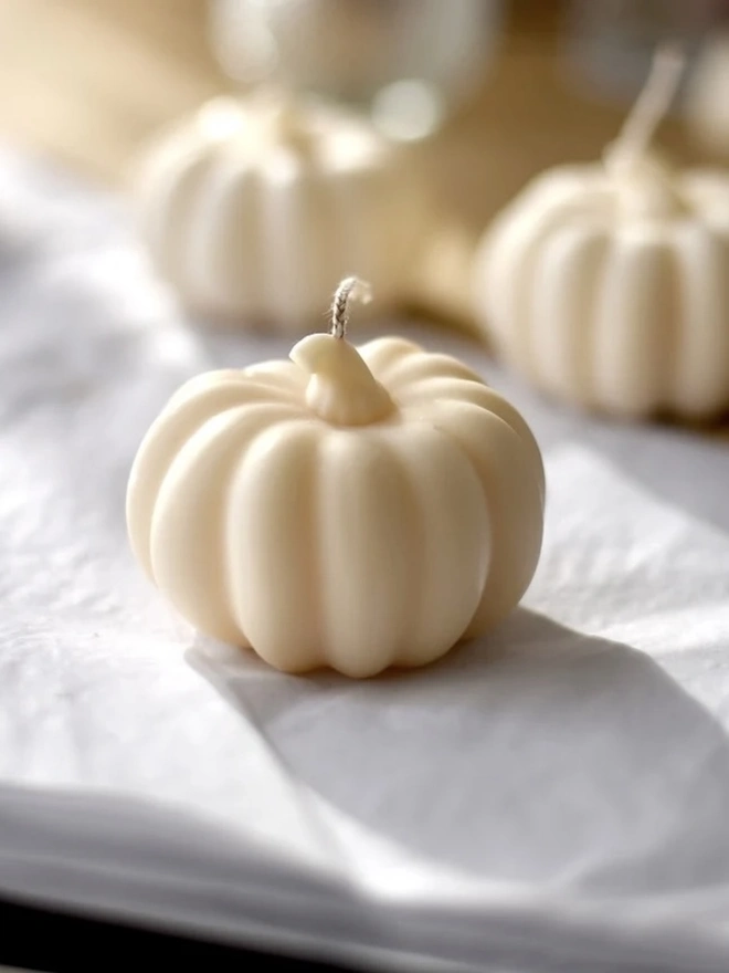 Medium Natural Wax Pumpkin Candle. Close-up of a small cream pumpkin candle with smooth ribbed sides and a curled stalk, resting on soft white linen. The minimalist design adds a subtle seasonal touch to shelves, mantelpieces and place settings.