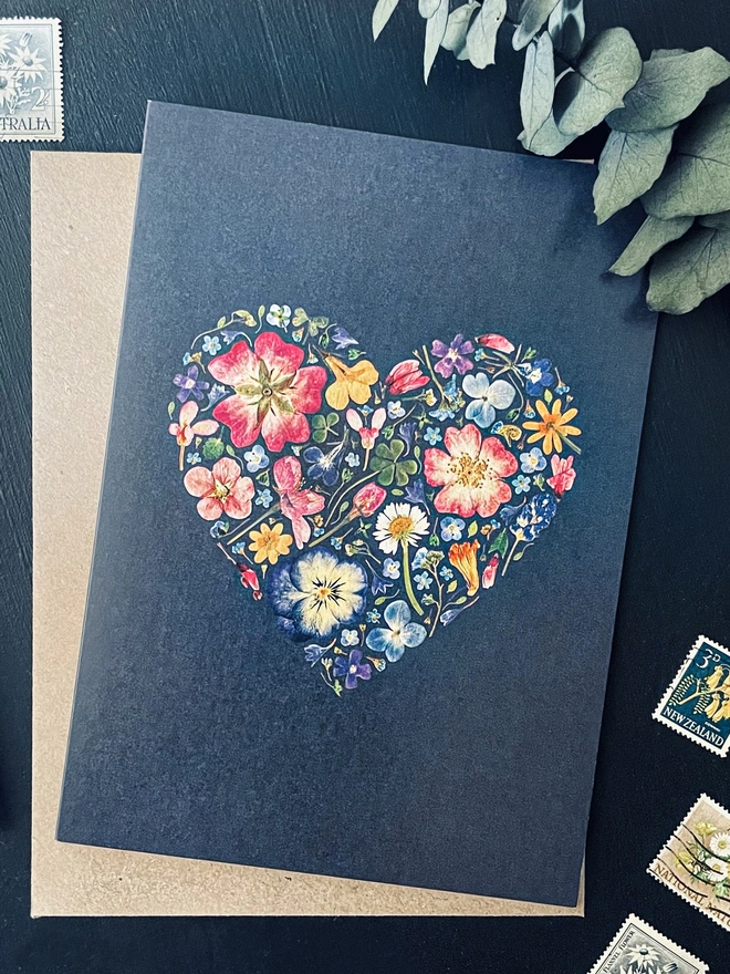 Close-Up of Inky Coloured Floral Greetings Card with Pressed Flower Heart Design - Brown Kraft Envelope - Dark Charcoal Coloured Desk - Postage Stamps, Silver Parker Pen, Dried Eucalyptus