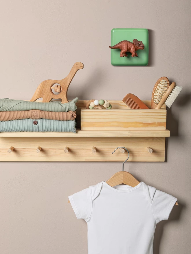 A dinosaur light switch on a dusky pink painted wall. There is a pine shelf with folded clothes on it, a wooden dinosaur rattle, wooden brushes and a white baby grow on a wooden hanger hanging from a line of pegs. The brown triceratops dinosaur on the light switch is used as the rotary dimmer switch knob. The light switch plate is beryl green and made of metal, epoxy coated steel. The triceratops is made of plastic. The brand is Candy Queen Designs.