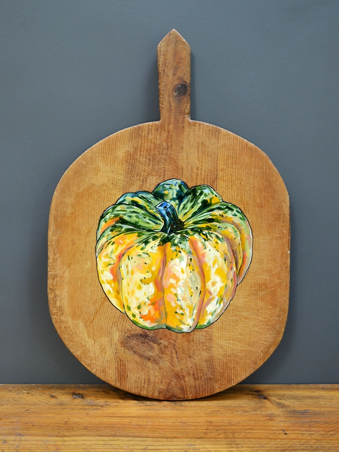 Wooden chopping board with handpainted design of a carnival squash standing against a wall