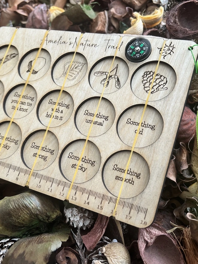 A personalised wooden nature scavenger hunt board displayed on an outdoor surface. The board is rectangular with rounded corners and engraved with 15 small collection slots held by clear bands. A custom name is engraved at the top, and a button compass with a wrist lanyard is attached for carrying. The wood has a smooth, light finish, and no items are placed in the slots; only the board is shown.