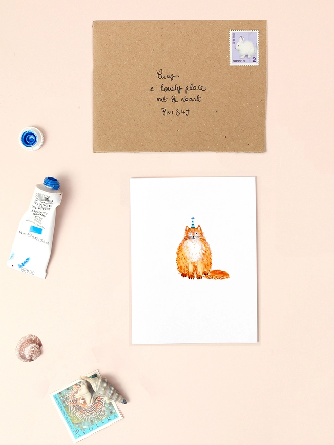 An image of a mini, rectangular white greetings card with an illustration of a big fluffy ginger cat wearing a party hat with a bushy tail. The mini greetings card is placed on top of a rectangular brown kraft envelope.