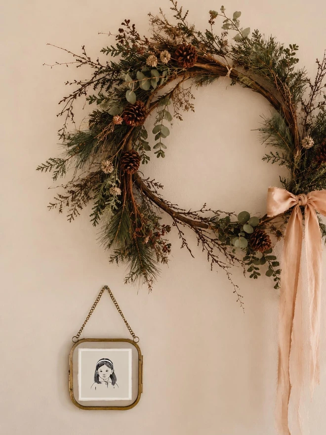 a miniture hand drawn portrait of a little girl framed in a sweet glass and brass frame hung on a wall next to a christmas wreath