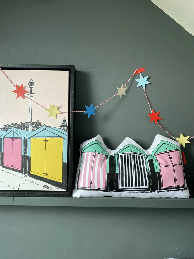 Row of beach huts handmade cushion by the artist Flo Snook. A pink and sea-green cushion in the shape of three beach huts in a row, on a shelf next to an original painting of beach huts by Flo Snook, with a colourful star garland hanging over them