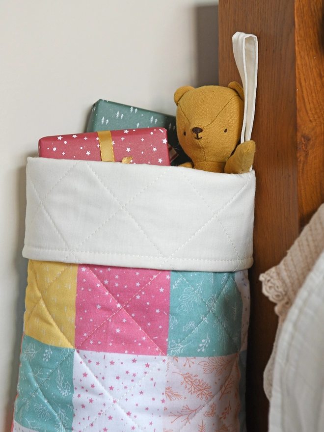 A quilted patchwork stocking in various pastel colours stands beside a wooden bed with ivory bedding. Small gifts and toys poke out of the top.