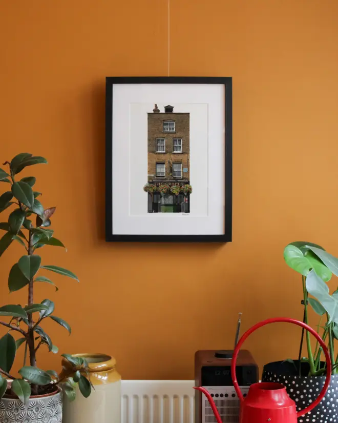 Limehouse The Grapes Giclée Print in black frame hanging on mustard yellow in room with plants and pots