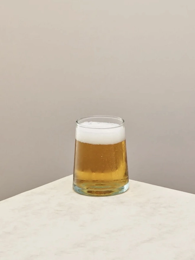 Tumbler Glasses (Set Of 1 Or 4), a tumbler glass sitting on a plain counter top against a plain backdrop. The glass contains a beer in it. 