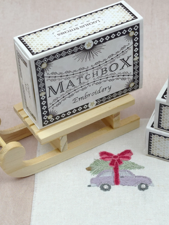 An embroidered lilac purple car carrying a Christmas fir tree on the roof, strapped in with a giant pink bow. Alongside the Matchbox displayed on a little wooden sleigh.