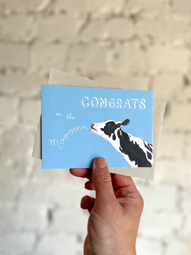a sky blue card with an illustration of a black and white cow and the text "congrats on the moooove", with a light grey envelope