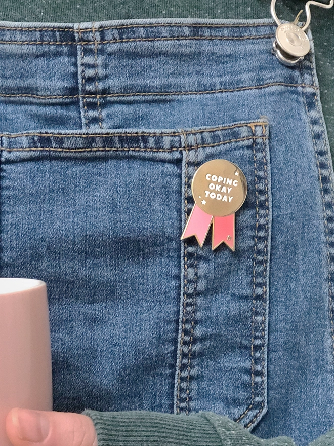 A pink and gold enamel pin badge in the shape of a rosette is pinned to blue dungarees. It has the words "Coping okay today".