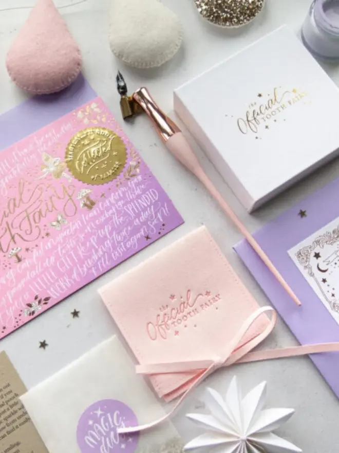 The pink-lilac notecard with its gold seal sits beside a white gift box and blush pink pouch. Felt teardrop decorations and a rose gold dip pen add to the scene.