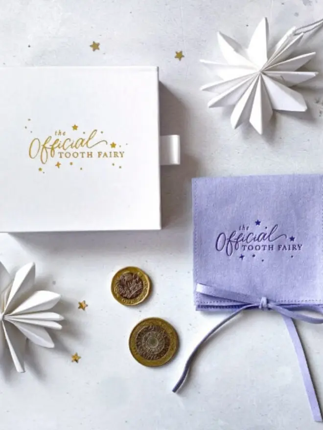 A white gift box with the gold foil "The Official Tooth Fairy" logo sits beside a lilac suede pouch tied with a bow. Two pound coins and white paper stars are scattered around them.