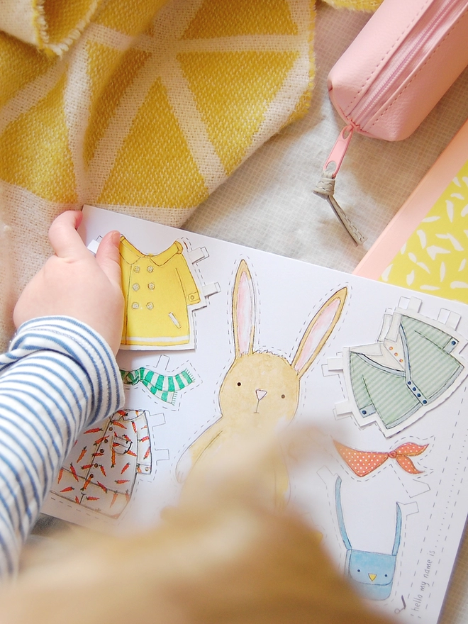 A child is playing with a dress up rabbit paper doll that has been cut from an illustrated greetings card.