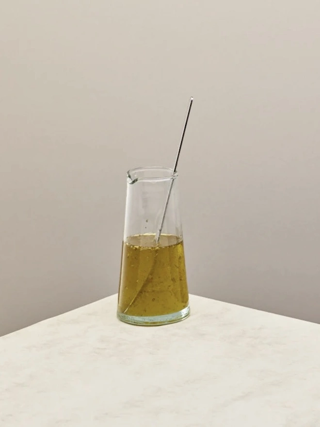 Glass Jugs (Set Of 2), a large glass jar sitting on a plain counter top against a grey backdrop. It is filled with oil. 
