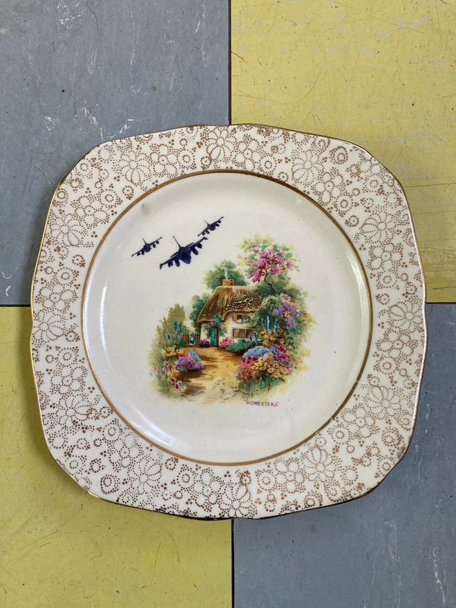 A gold and white china plate featuring a cottage with fighter jets in the sky. A gold and white china plate featuring a cottage with fighter jets in the sky.