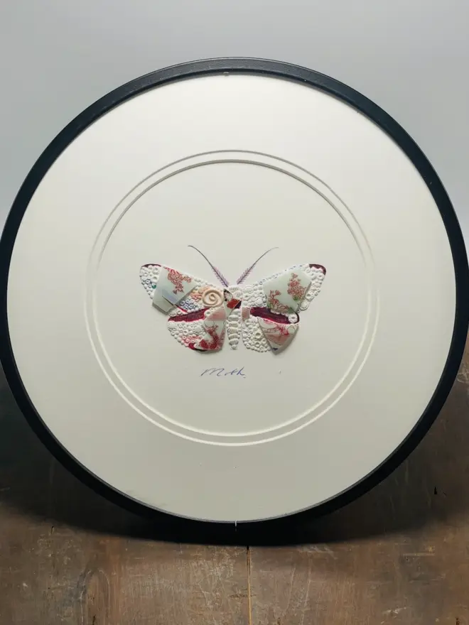 Hand-drawn, painted, mosaic clay, vintage pink floral Geisha pottery insect on white double mounted card, in a black circular frame.