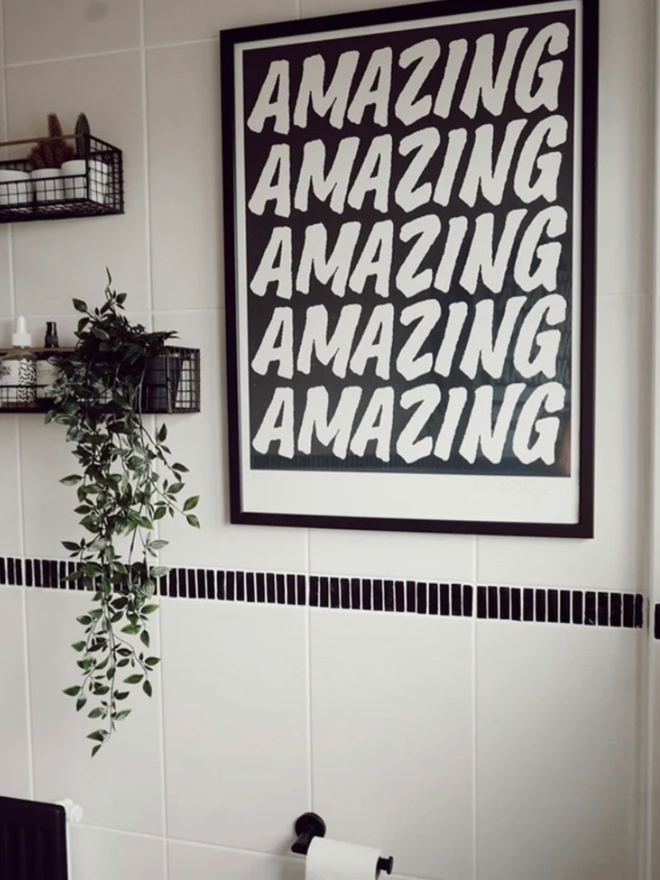 Black Amazing Poster. A framed print hanging against a tiled wall. The print has a black background and AMAZING written in a simple, all caps font five times.