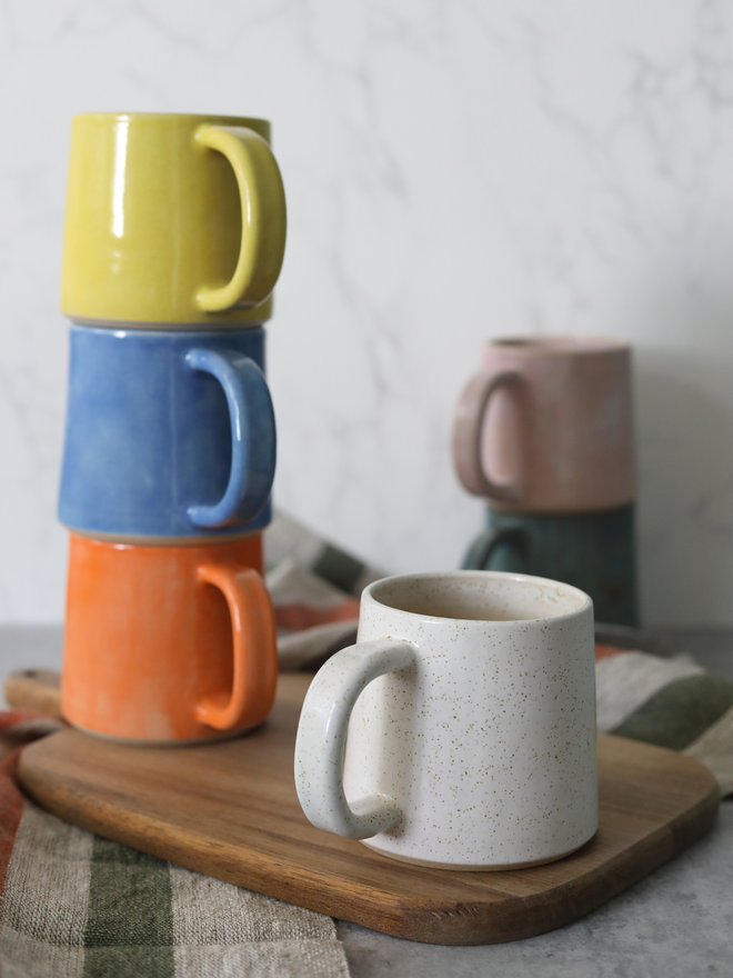 Several handmade ceramic standard mugs styled on a wooden board — speckled white in the foreground, with yellow, blue, and mandarin orange stacked behind, and blush pink and teal mugs softly blurred in the background, styled with linen cloths against a white marble background