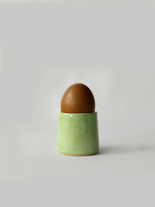 A handmade ceramic egg cup in spring green glaze holding a brown egg, photographed against a plain white background
