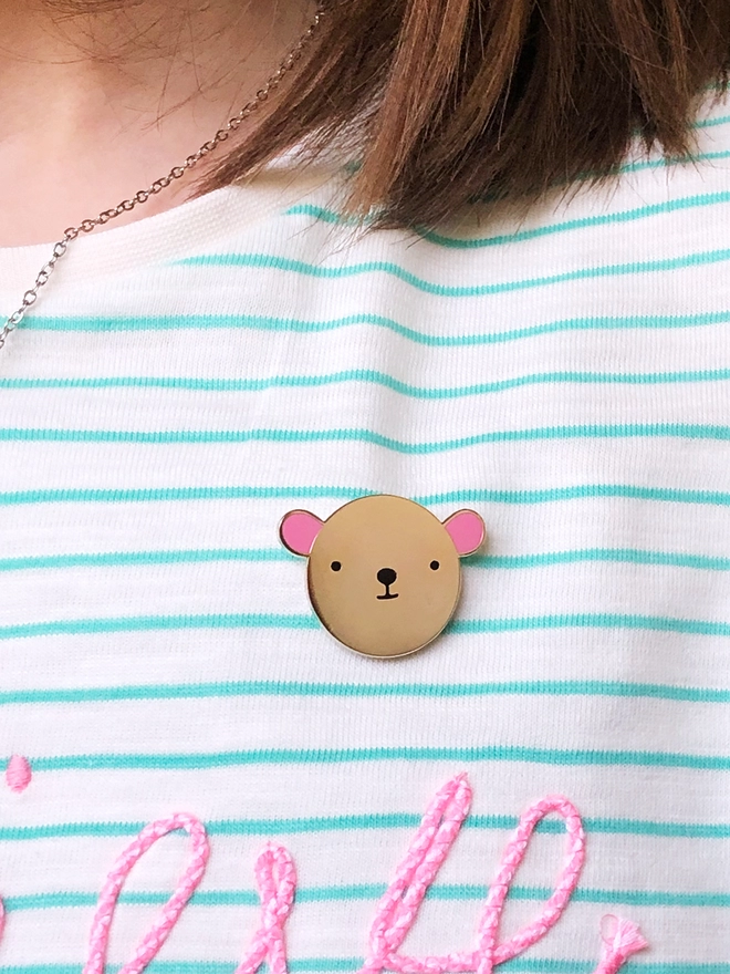 A gold bear pin with pink ears is pinned on a white and green striped top.