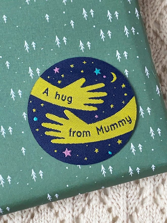 A navy blue and yellow woven patch is laying on a wrapped gift. The design is two arms in a hug shape with the words "A hug from Mummy" along them. 