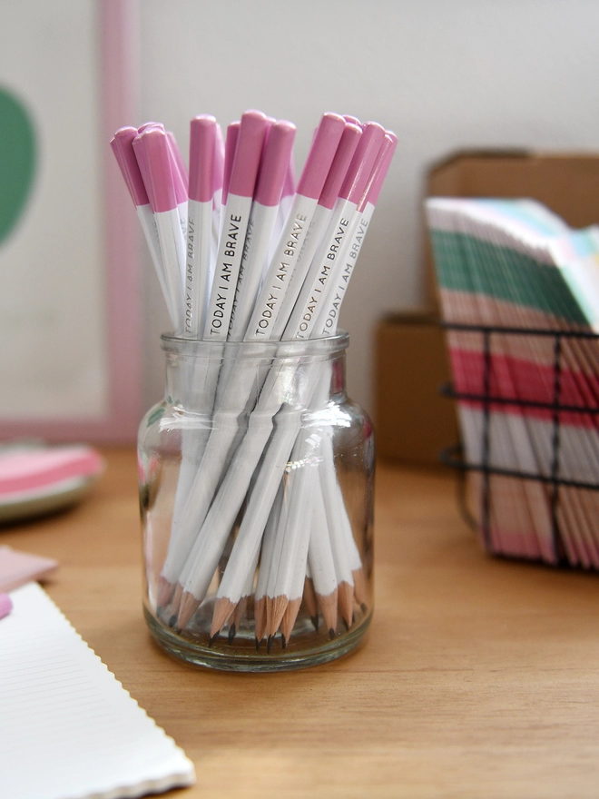 A glass jar full of white pencils with pink tips stands on a wooden desk surrounded by various stationery items.