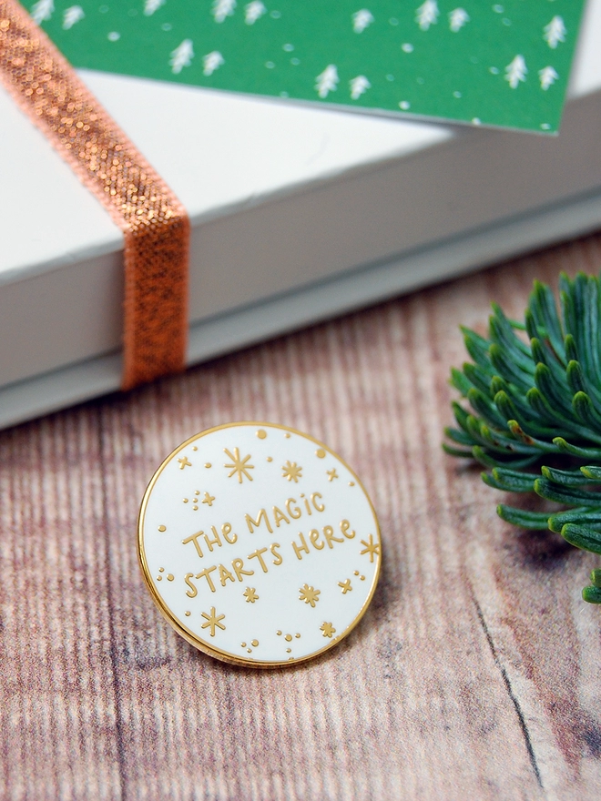 A white and gold enamel pin badge with a starry design and the words "The magic starts here" is beside a gift box on a wooden table.