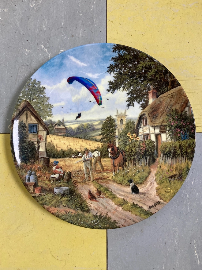 A vintage plate featuring farm workers with a modern day paraglider about to land in the field.