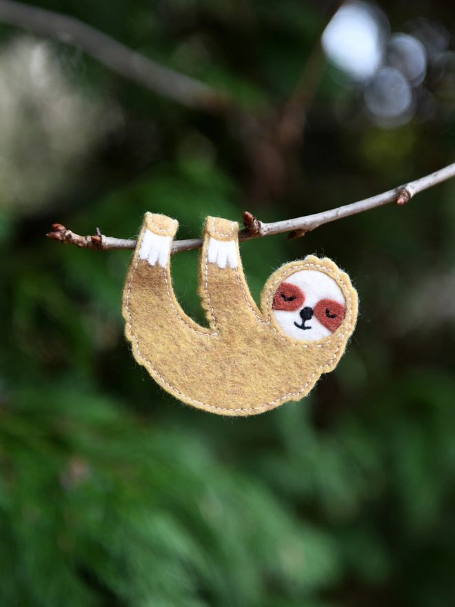 A handmade sloth finger puppet made from beige and white felt is hanging from a tree branch.