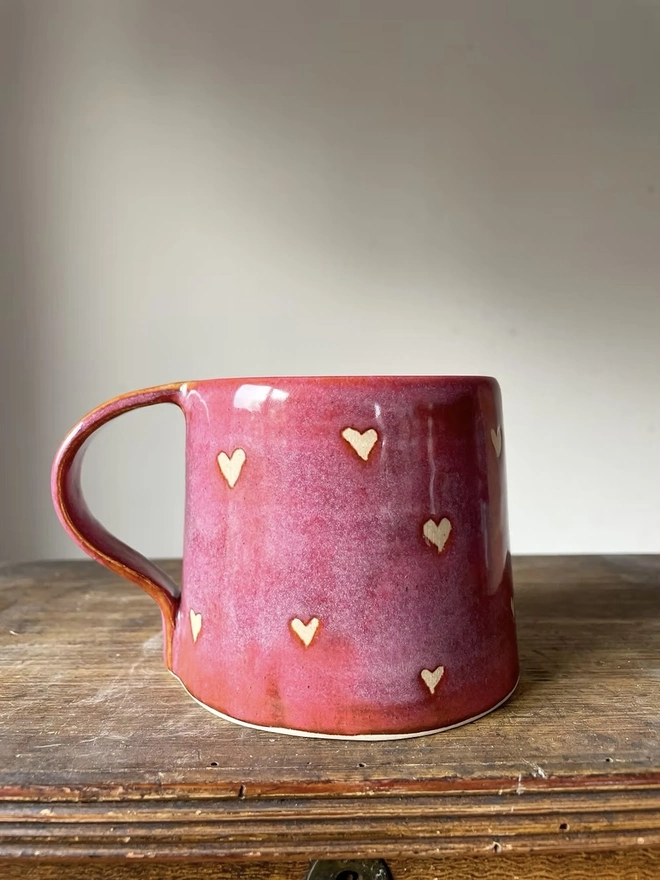 Love Heart Mug Raspberry , a red heart mug on a wooden surface in front of a plain background. 