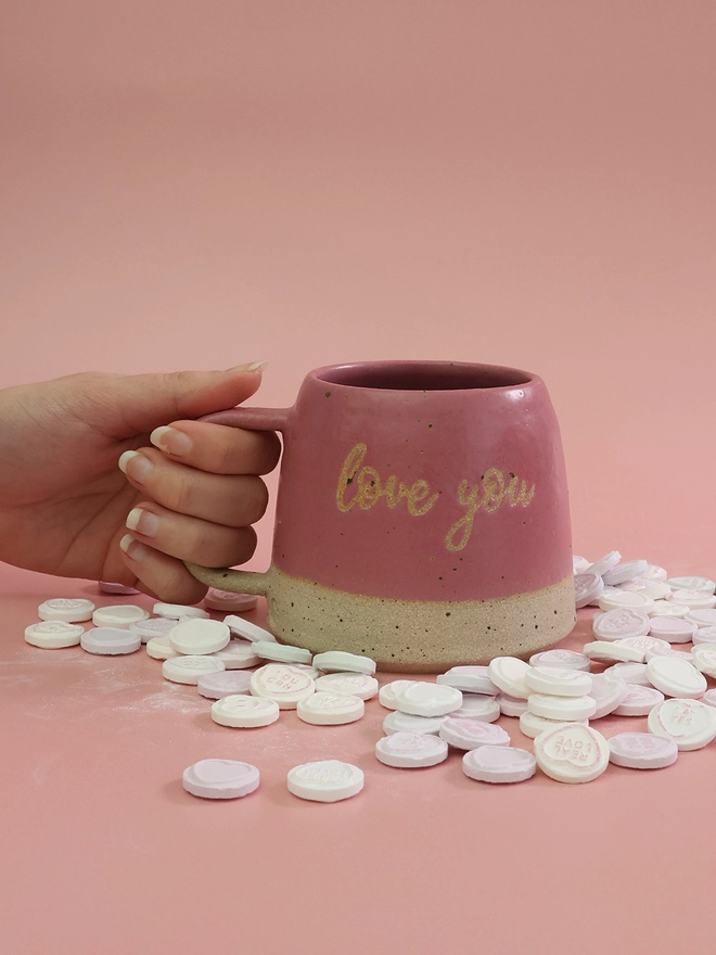 a hand is holding a handmade bright pink mug with the phrase love you on the body of the mug, it sits on a pink background surrounded by love heart sweets 