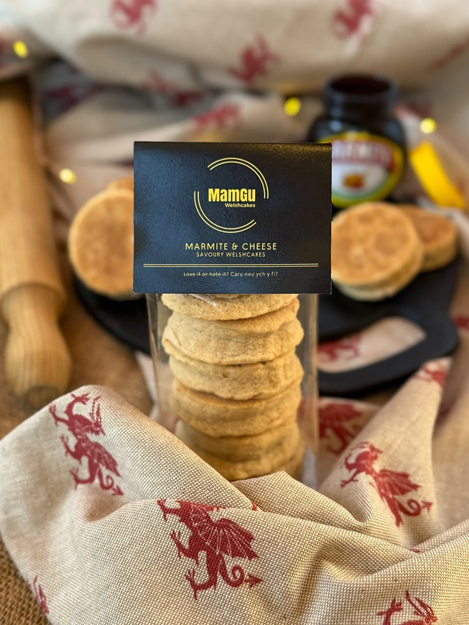 6 marmite and cheese welsh cakes stacked in packaging with a black MamGu label and a messy rustic kitchen 