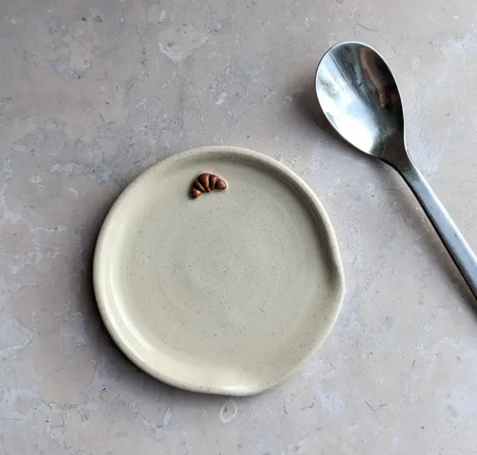 Stoneware spoon rest in vanilla bean glaze with a croissant design and a spoon beside it, on a grey marble background.