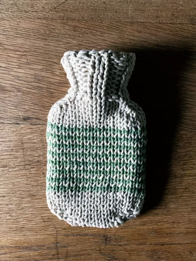 Mini Hot Water Bottle in hand knit string cover with green stripe on an oak table