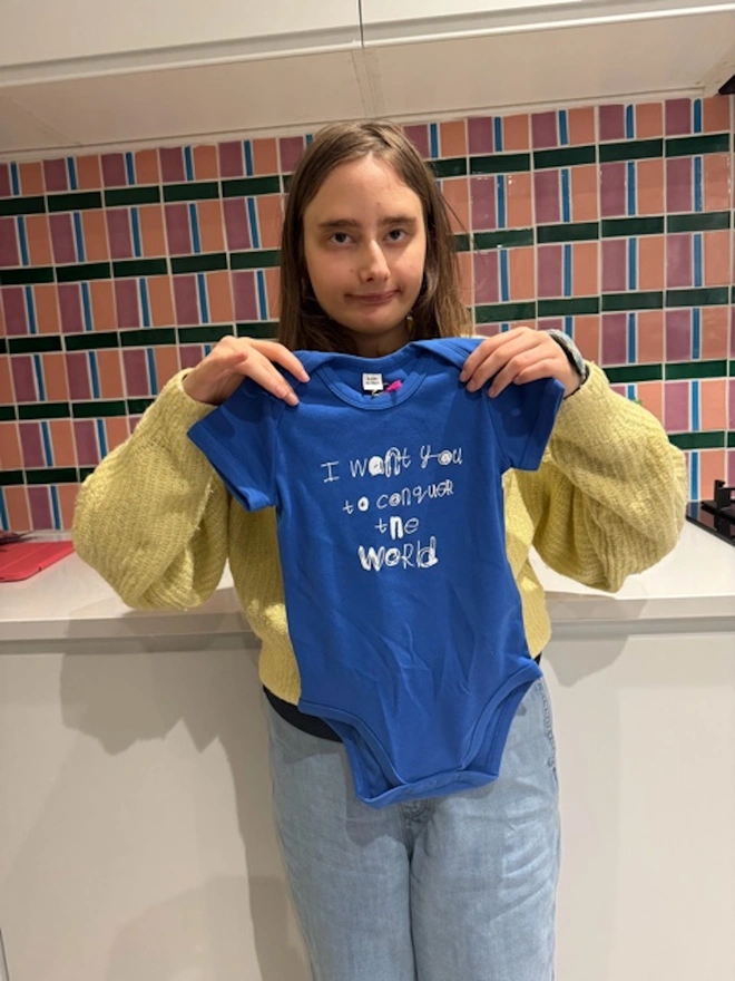 Piper holding up a blue baby grow with her unique white font on the tummy