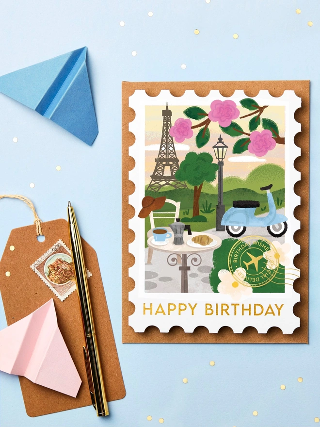 Stamp-shaped greeting card featuring an illustration of Paris. The card is on a blue background with paper aeroplanes and stationery around it.