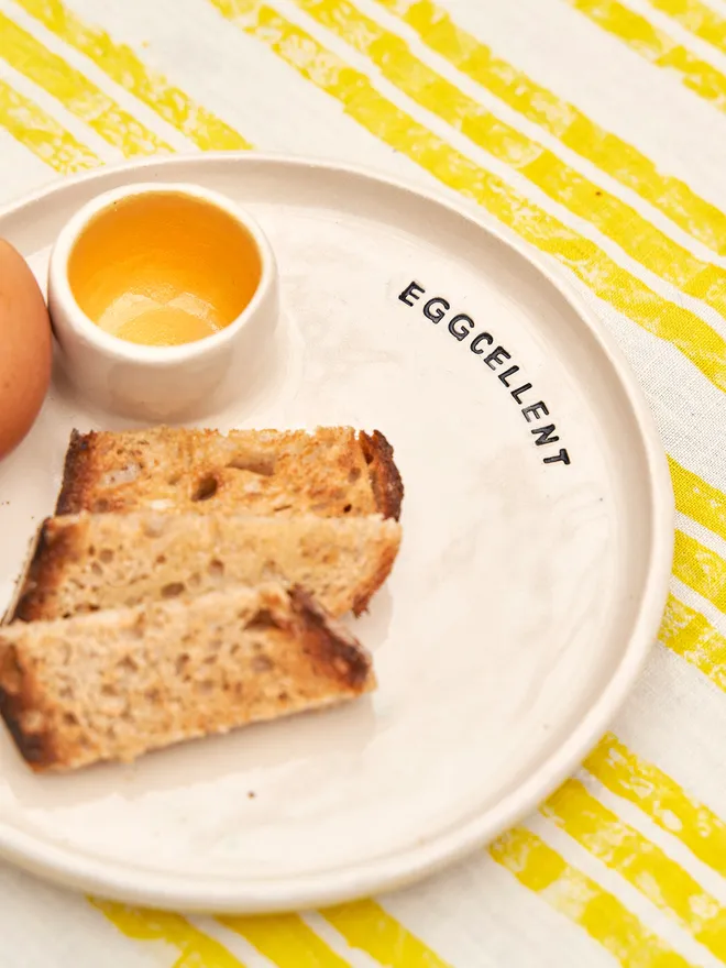 Egg And Soldier Plates, a plate with an integrated egg cup placed on a yellow striped surface. The wording reads 'eggcellent' 