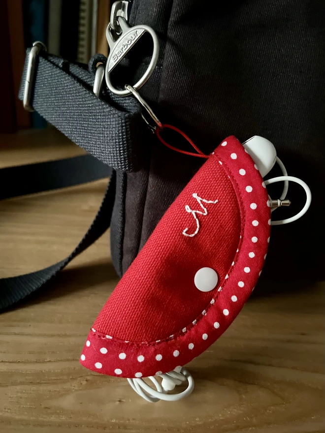 Soft red cotton earphone case embroidered with initial, polka dot trim and snap closure., holding earphones, attached to bag.