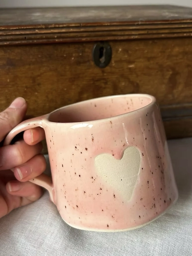 Heart Mug Strawberry Pink, person holding pink mug by handle in front of a wooden background. 