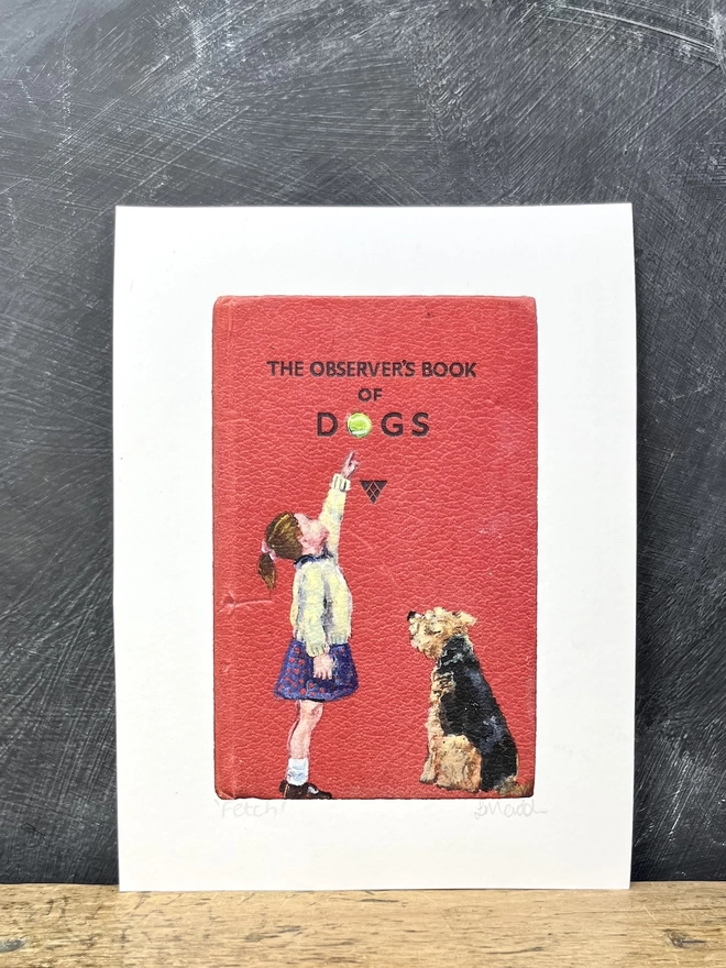a print propped against a blackboard showing an aged observer book cover of dogs with a painting of a little girl pointing to a tennis ball with an airedale or welsh terrier looking up