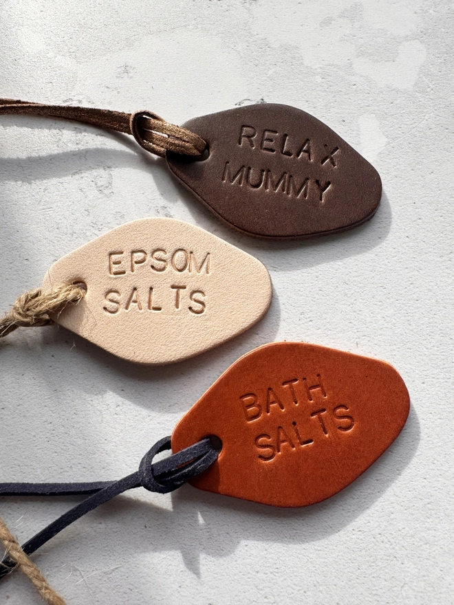 Three hand-stamped leather tags in brown, cream, and tan, with the words 'RELAX MUMMY,' 'EPSOM SALTS,' and 'BATH SALTS' embossed. Each tag has a cord for tying around jars, displayed on a light, textured surface