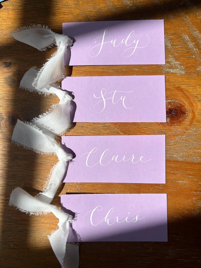 Personalised Calligraphy Wedding Lilac Place Cards, lilac place cards with a white ribbon grouped together on a table. 