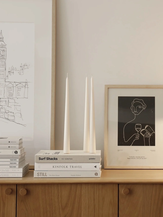 White Stem Candles (Set Of 3). Three unlit cream taper candles balanced on a stack of neutral books in front of a light wood frame. The sleek freestanding shape needs no holder, which makes it easy to style on shelves and sideboards.